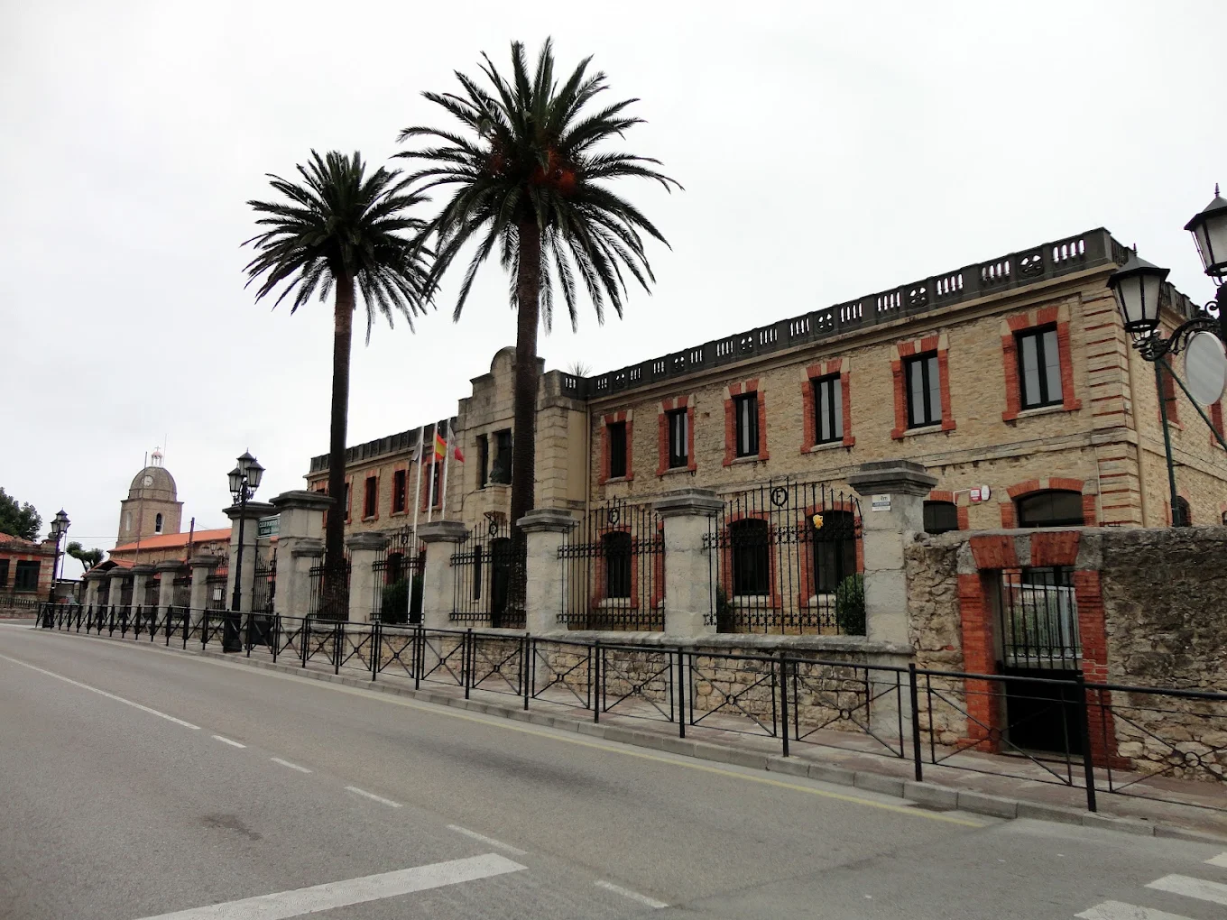 Vista general actual del Antiguo Colegio de San José en Suances, con la verja de forja, las palmeras del acceso y la torre de la iglesia parroquial al fondo