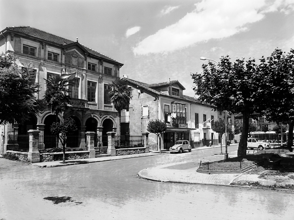Fotografía histórica en blanco y negro del Ayuntamiento de Suances en los años 70, con un coche de época junto al soportal sin acristalar