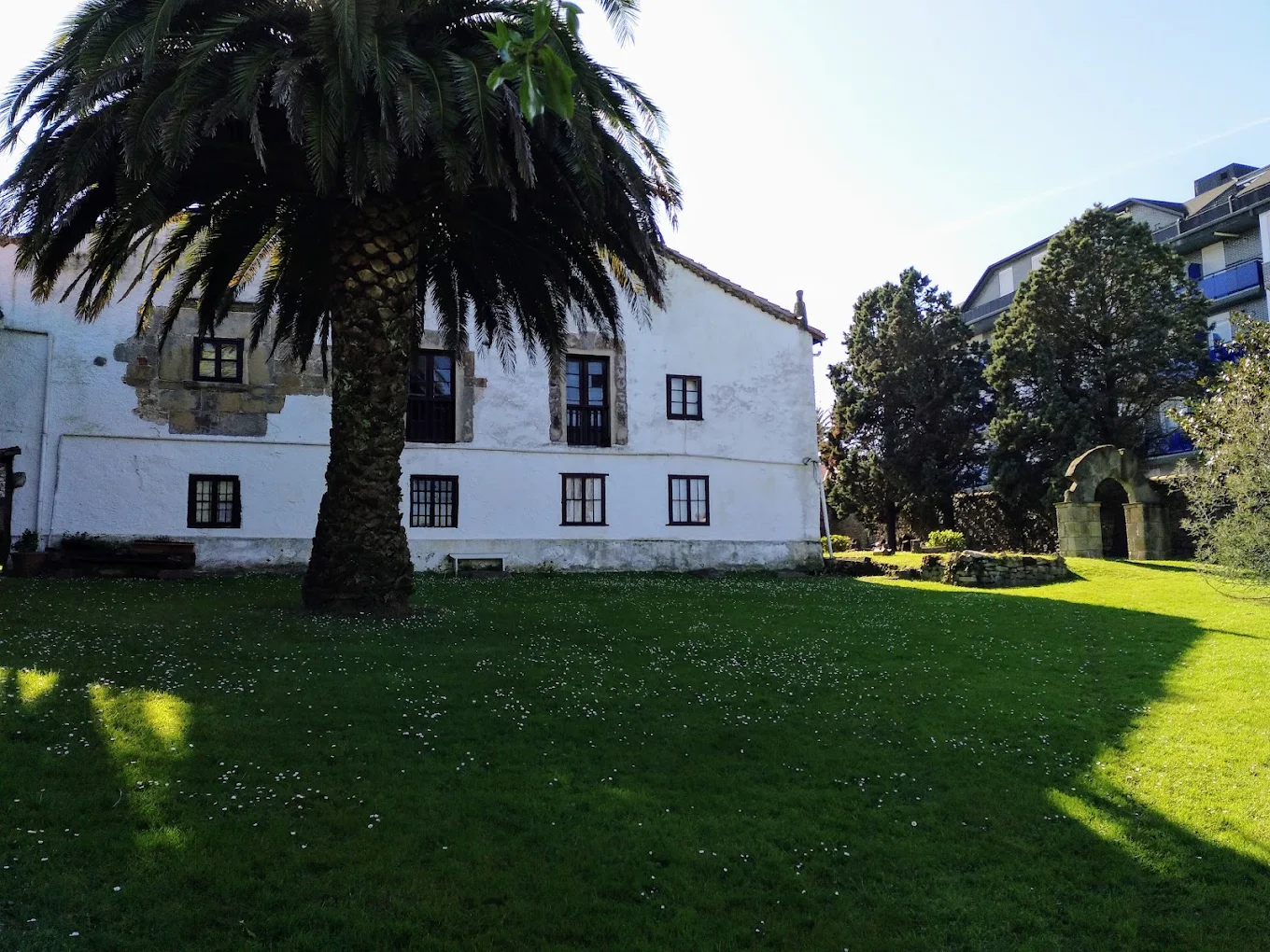 Ala trasera encalada de la Casa de los Polanco con una gran palmera y el jardín privado del recinto, con un arco de piedra al fondo