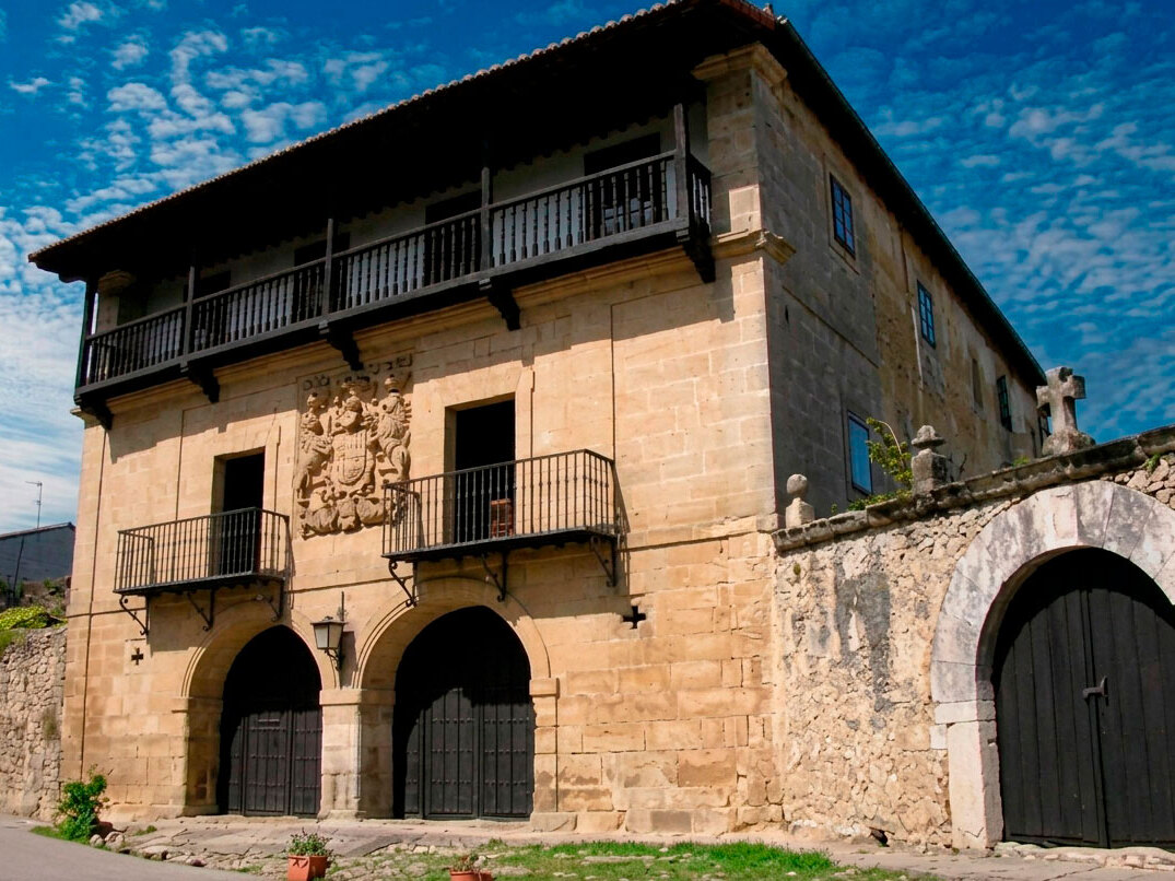 Vista lateral de la Casona de los Tagle en Santillana del Mar bajo un cielo azul, con el escudo entre los dos balcones y el muro de piedra del acceso