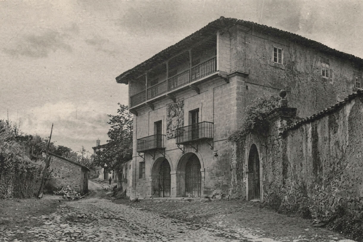 Fotografía antigua en blanco y negro de la Casona de los Tagle en Santillana del Mar, con la calle empedrada sin asfaltar y el escudo bajo el balcón