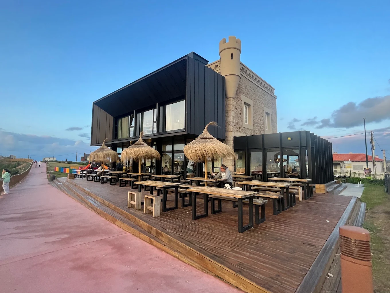 Terraza exterior del hotel El Castillo de los Locos al atardecer, con sombrillas de paja y la torre del castillo al fondo
