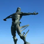 Escultura El Hombre de los Vientos en Suances Escultura El Hombre de los Vientos en Suances, figura de bronce con los brazos abiertos sobre fondo de cielo azul