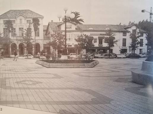 Foto antigua en blanco y negro de la plaza de Viares en Suances en los años 90, con el edificio del Ayuntamiento al fondo, farolas y coches de época aparcados alrededor del parterre central