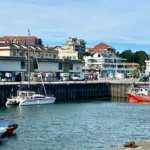 Vista general del puerto de Suances con velero y catamarán amarrados junto a un pesquero rojo y edificios del pueblo al fondo