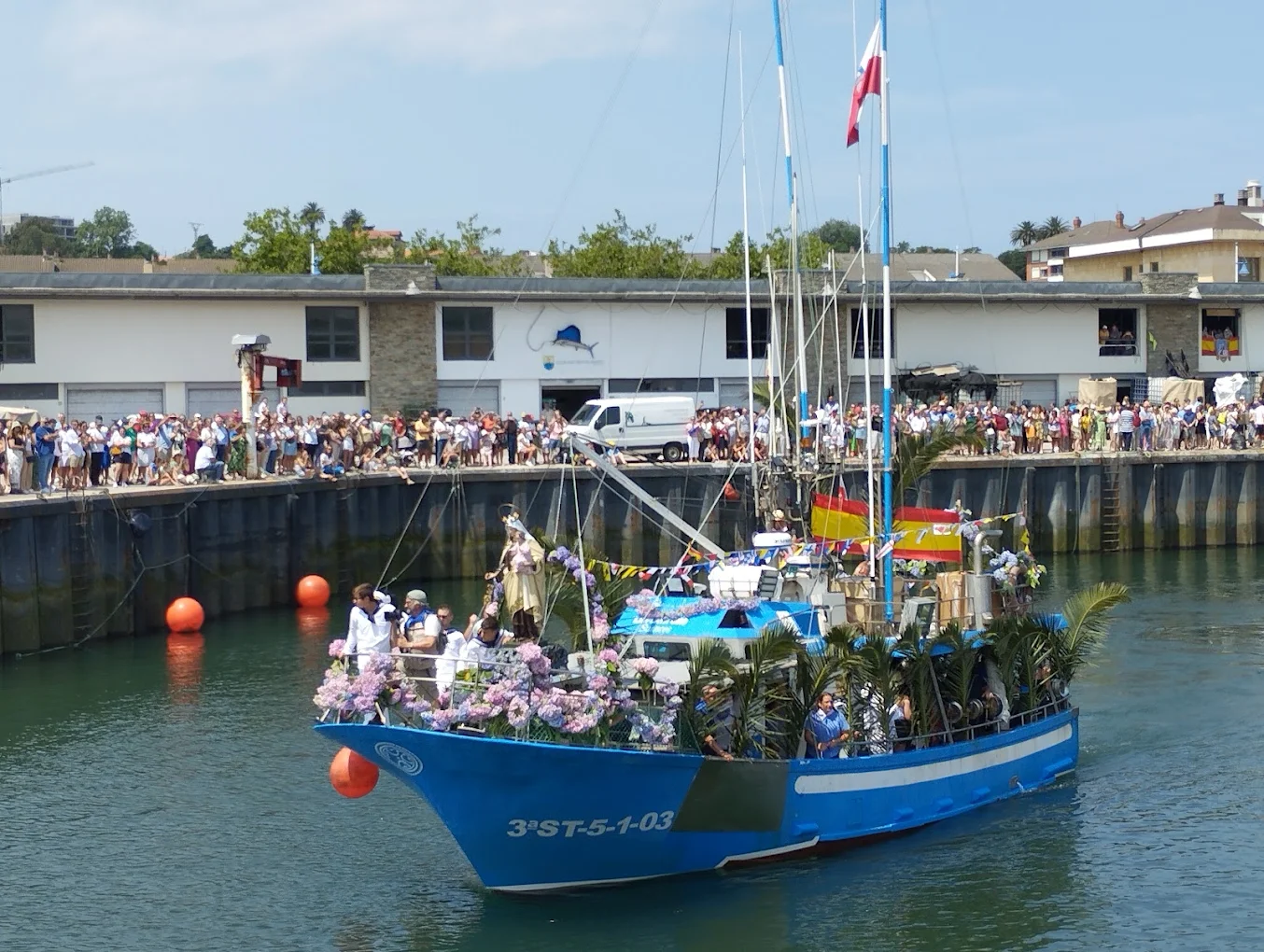 Procesión marítima de la Virgen del Carmen en el puerto de Suances con la embarcación engalanada de flores y numerosos vecinos en el muelle