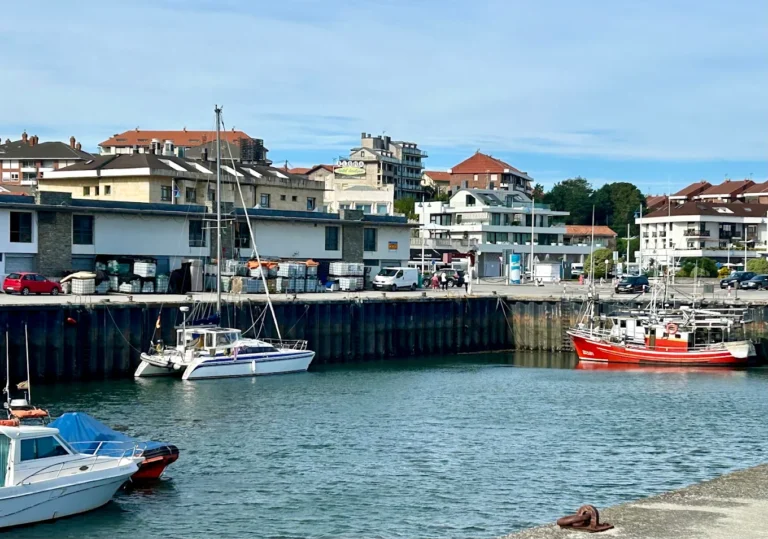 Vista general del puerto de Suances con velero y catamarán amarrados junto a un pesquero rojo y edificios del pueblo al fondo