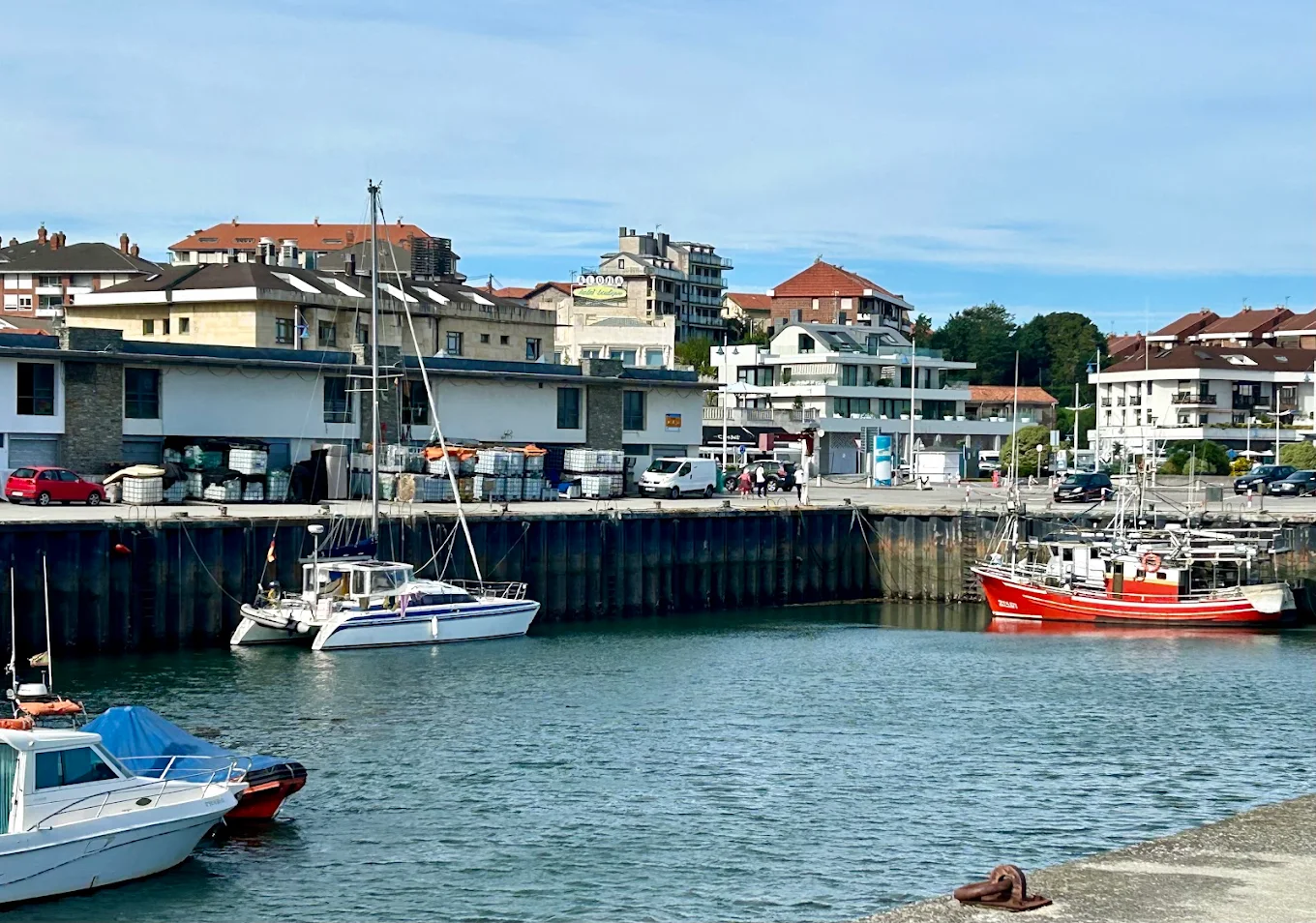 Vista general del puerto de Suances con velero y catamarán amarrados junto a un pesquero rojo y edificios del pueblo al fondo