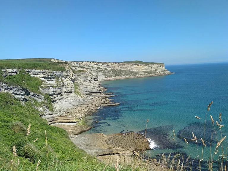 Acantilados escalonados de roca caliza en Punta Ballota (Tagle, Suances), con sus estratos horizontales asomando sobre una cala de aguas turquesas