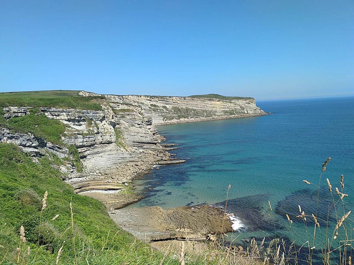 Acantilados escalonados de roca caliza en Punta Ballota (Tagle, Suances), con sus estratos horizontales asomando sobre una cala de aguas turquesas